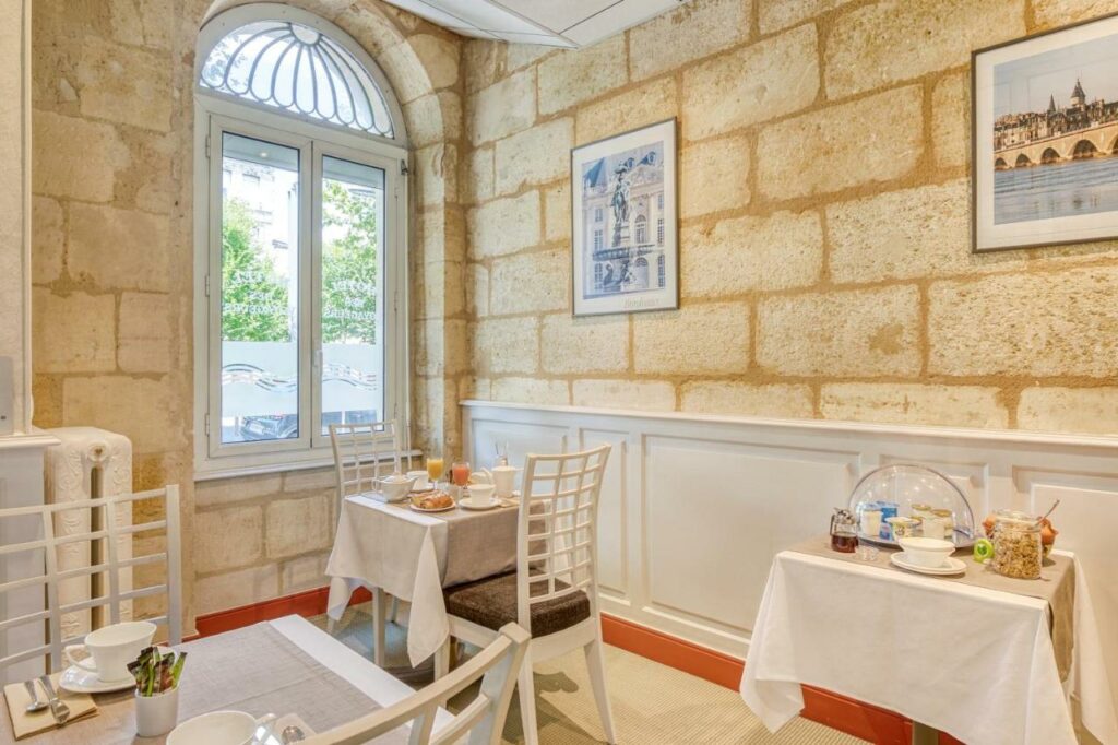 Área de refeições do Hôtel des Voyageurs Centre Bastide. Existem várias mesas pequenas  com café da manhã destribuidas pelo ambiente. Há também uma janela na parede.
