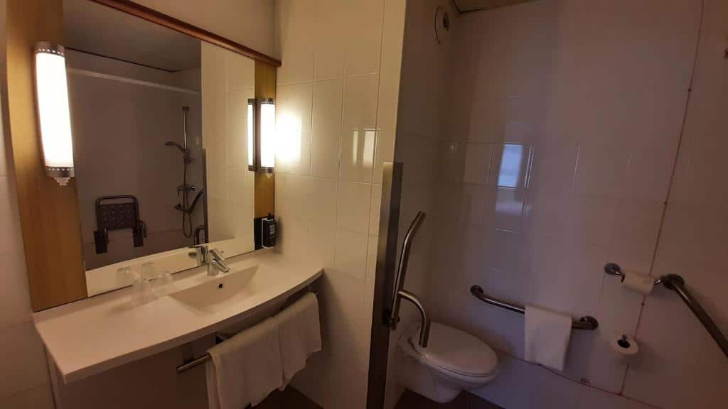 Banheiro adaptado do Ibis Lisboa José Malhoa com barras de apoio, espelho, pia mais baixa, box sem vidro e com cadeira para sentar-se
