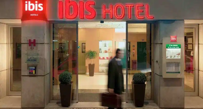 Entrada do Hotel ibis Lisboa Saldanha com placas vermelhas e um homem saindo do local segurando uma bolsa, para representar hotéis Ibis em Lisboa.