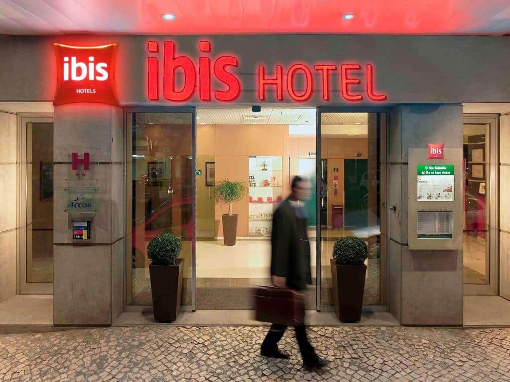 Entrada do Hotel ibis Lisboa Saldanha com placas vermelhas e um homem saindo do local segurando uma bolsa, para representar hotéis Ibis em Lisboa