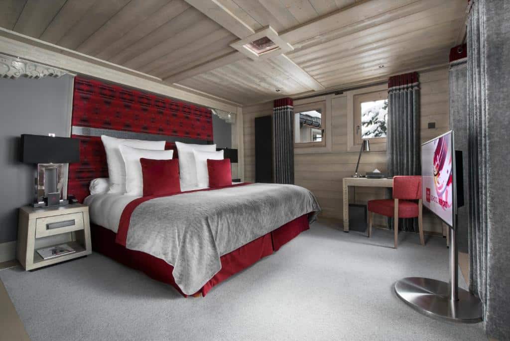 Quarto fo Hotel Le K2 Altitude com uma cama de casal, uma mesinha ao ladoo, uma televisão logo em frente e uma mesa e cadeira encostadas em uma parede com duas janelas. Foto para ilustrar post sobre hotéis em Courchevel.