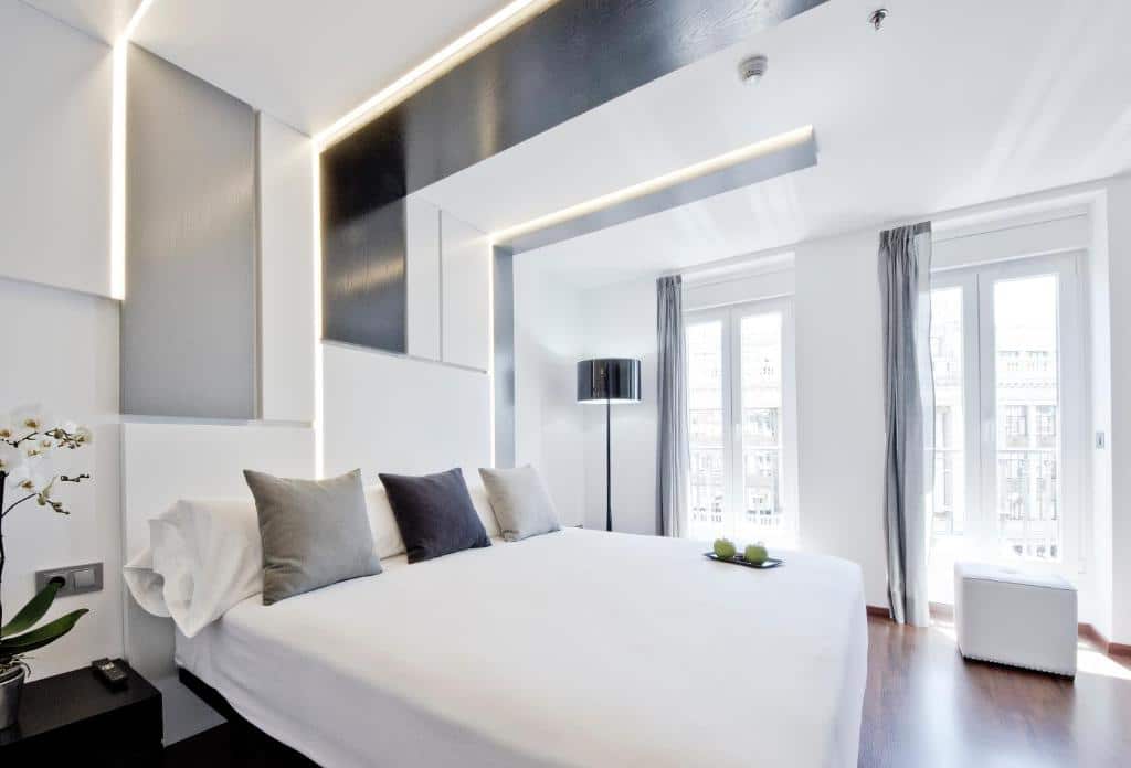 Quarto do Hotel Regina, uma das recomendações de hotéis em Madri. A cama fica no centro do quarto e tem mesinhas de cabeceira dos dois lado. A parede ao lado tem duas grandes janelas com cortinas, e de um lado fica um abajur enquanto do outro há um pufe. O local é decorado em estilo futurista.