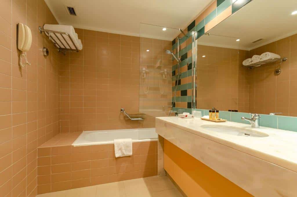 Banheiro do Hotel Roma com uma banheira, um espelho amplo com uma pia ampla embaixo, há toalhas brancas sob uma prateleira, para representar hotéis perto do Aeroporto de Lisboa