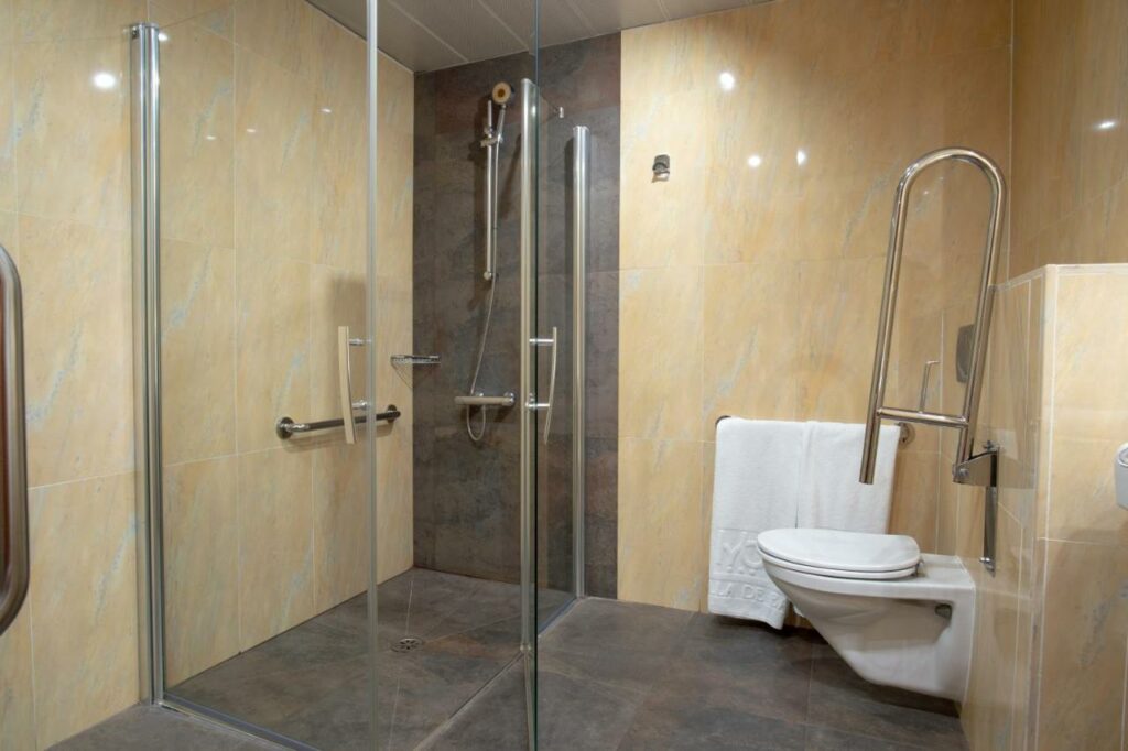 Banheiro acessível para PCD no Hotel Villa de Barajas. O box de vidro com chuveiro tem barras de apoio e, ao lado, uma privada branca também conta com as barras.