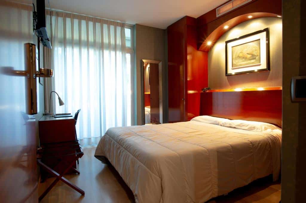 Quarto do Hotel Villa de Barajas, uma das recomendações de hotéis perto do aeroporto de Madri. A cama de casal tem um grande móvel ao redor com iluminação e um quadrinho, e encara uma mesa de trabalho com cadeira e luminária. Há uma TV na parede acima. Ao fundo fica uma janela com cortinas brancas e um espelho ao lado.