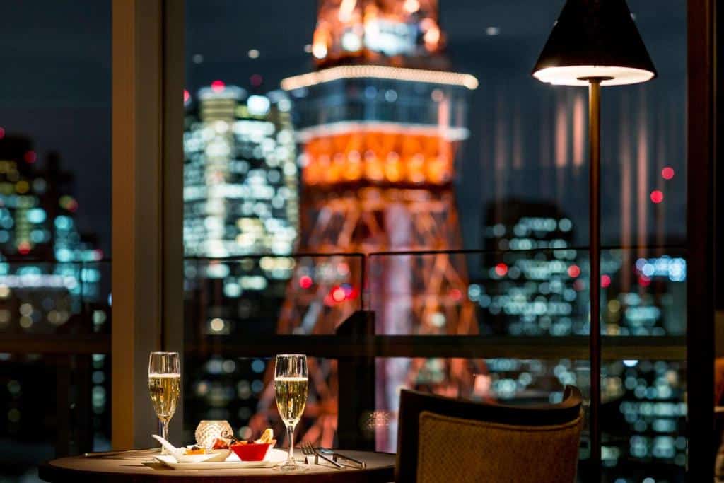 Foto profissional tirada de uma mesa de jantar no The Prince Park Tower. Vemos duas taças de champagne, um prato com entradas e uma pequena luminária na mesa. De frente, há uma janela grande com vista para a Torre de Tóquio.