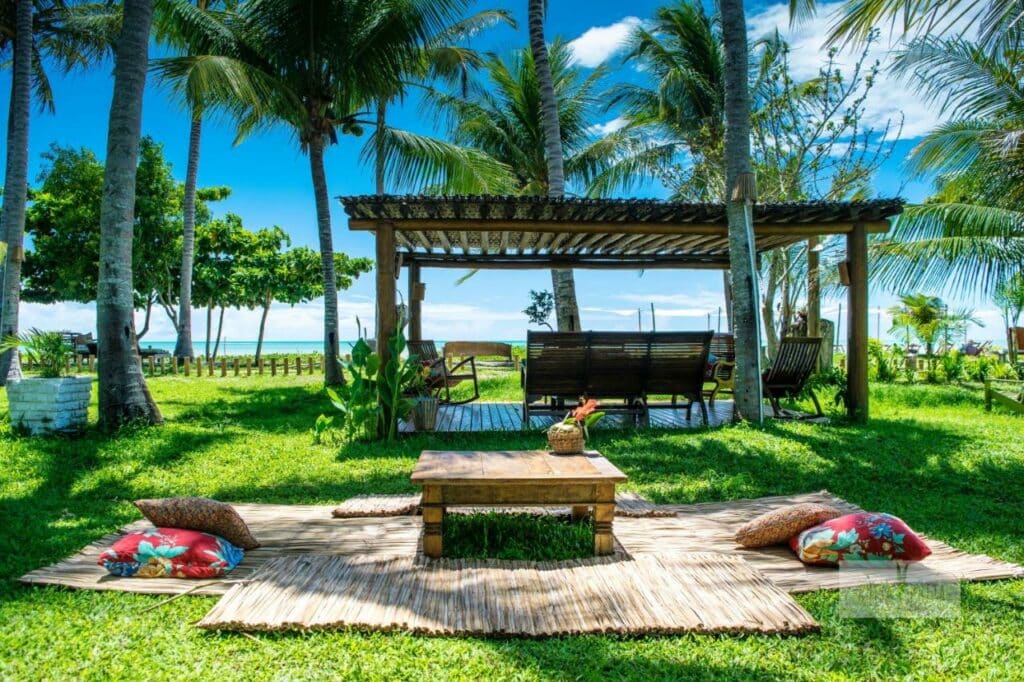 Área externa da Pousada Areia Preta. Uma mesinha no centro e esteiras ao redor com almofadas. Atrás cadeiras de descanso viradas para o mar. Grama, muitas palmeiras e atrás o mar.