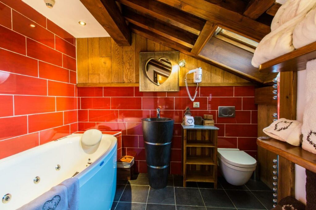 Bnaheiro do hotel Les Monts Charvin. Suas paredes são vermelhas, há um espelho em formato de coração pendurado na parede, um vaso sanitário bem baixo, uma prateleira cheia ade toalhas encostada na parede. Do lado oposto está a banheira.