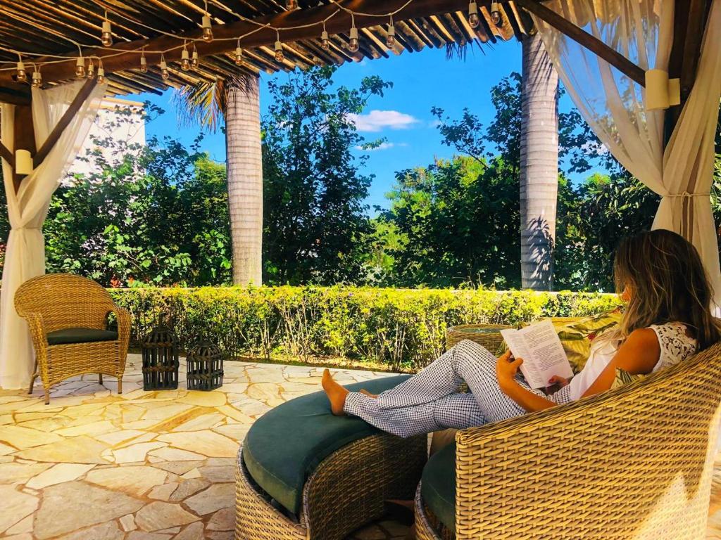 Imagem de uma área de descanso com cobertura da LS Villas Hotel & Spa. Há uma moça loira de costas para a câmera, sentada em uma poltrona lendo. No fundo podemos ver árvores, arbustos e um céu bem azul. Imagem utilizada para representar o post sobre pousadas em Águas de São Pedro.