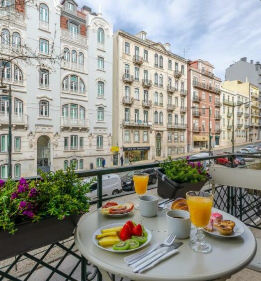Varanda de um dos quartos do LX51 Studios & Suites – Lisbon center com vista para uma rua com prédios antigos, na varanda há uma mesa com um café da manhã sob ela, para representar hotéis baratos em Lisboa