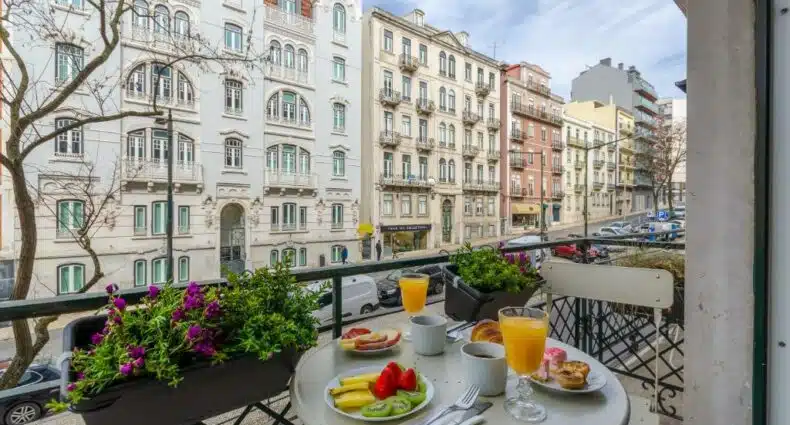 Varanda de um dos quartos do LX51 Studios & Suites – Lisbon center com vista para uma rua com prédios antigos, na varanda há uma mesa com um café da manhã sob ela, para representar hotéis baratos em Lisboa