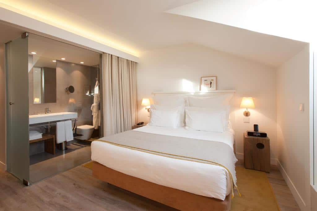 Quarto do Memmo Alfama – Design Hotels com uma cama de casal, logo ao lado, separado por um cortina, há uma banheiro com pia, vaso, roupões e espelho, do lado direito do cômoda há uma janela