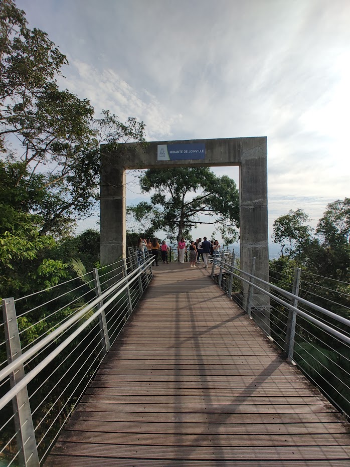 caminho de madeira, centralizado na imagem, que termina num arco quadrangular de concreto com os dizeres "Mirante de Joinville" escritos numa placa azul e preso ao arco, na parte de cima. Ao fundo há várias pessoas admirando a paisagem.