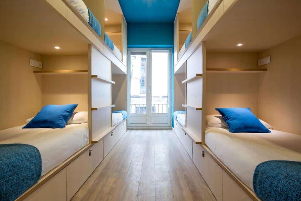 Quarto do Mola Hostel. As beliches estão dos dois lados do quarto e possuem cobertor, travesseiro e almofada. O corredor entre as camas leva a uma grande janela de vidro.