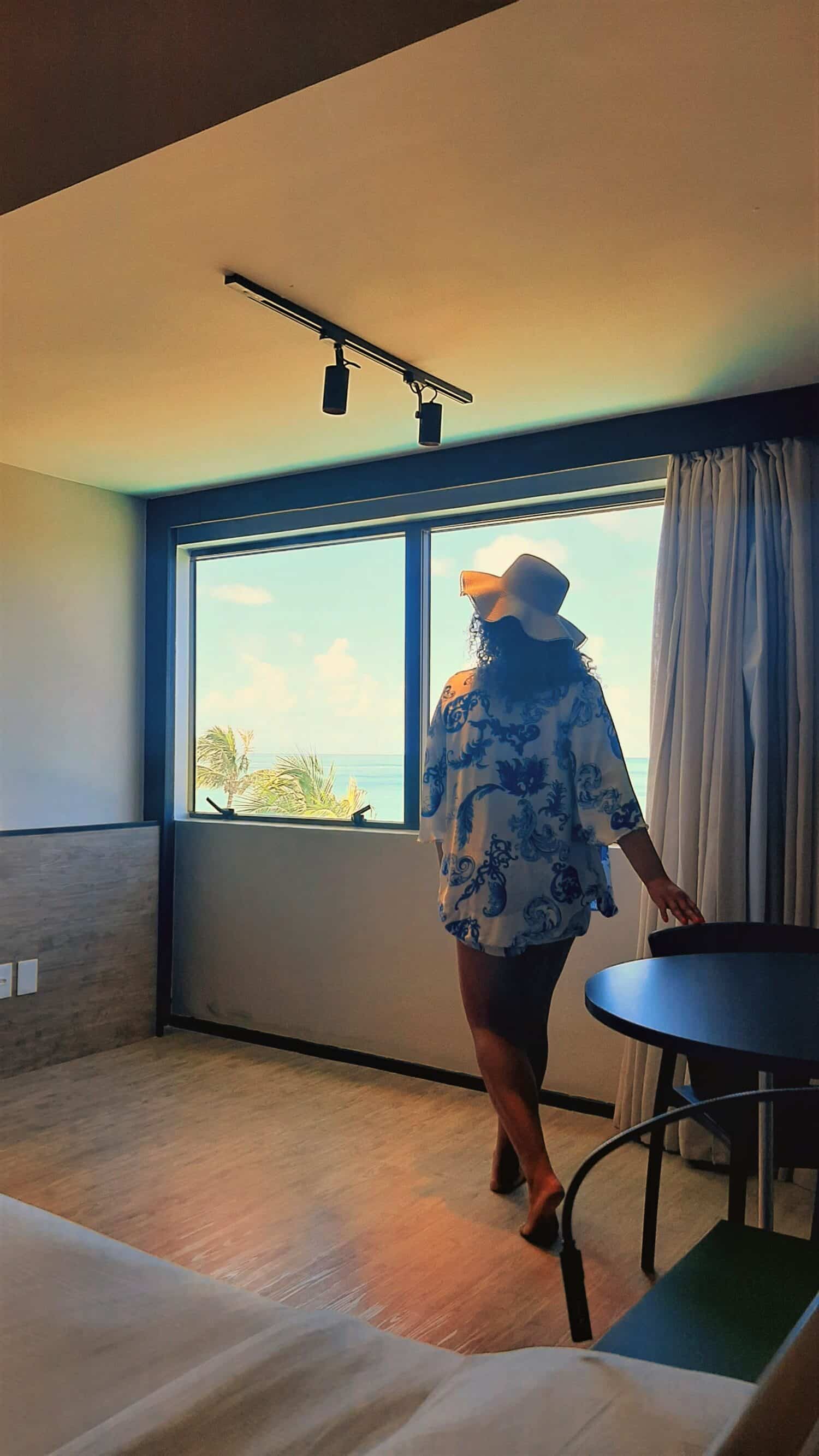 Mulher de costas, com o cabelo solto e chápeu de praia, dentro de um dos quartos do Ritz Lagoa da Anta olhando para a janela ampla com vista do mar.