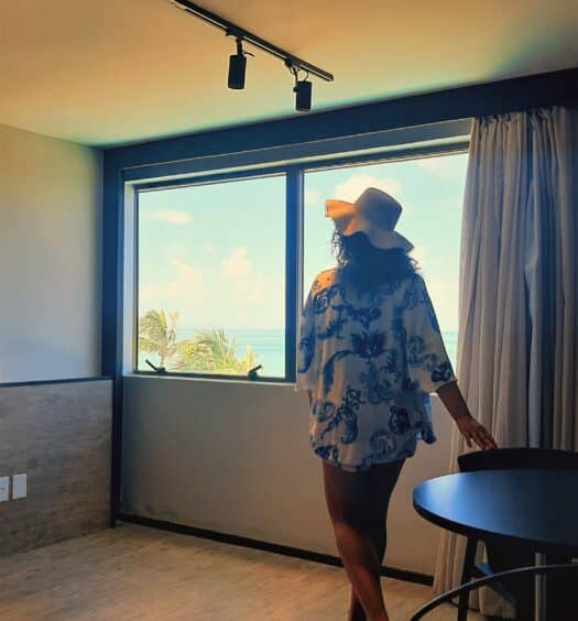 Mulher de costas, com o cabelo solto e chápeu de praia, dentro de um dos quartos do Ritz Lagoa da Anta olhando para a janela ampla com vista do mar.