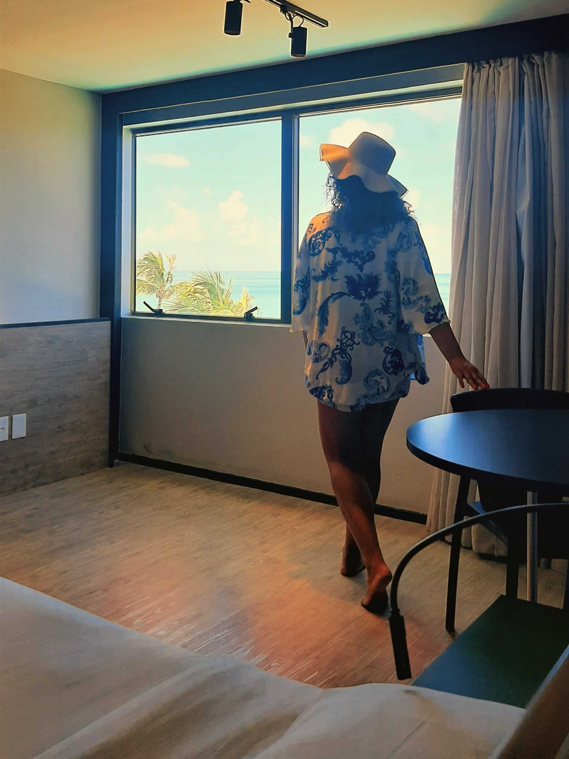 Mulher dentro de um quarto do hotel Ritz Lagoa da Anta, em Maceió, vestida de roupa branca com estampas azuis e chapeu, de costas, com uma das mãos em uma cadeira de uma mesa redonda olhando para uma janela ampla de vidro com vista do mar e folhas de uma árvore. Aparece também parte da cama