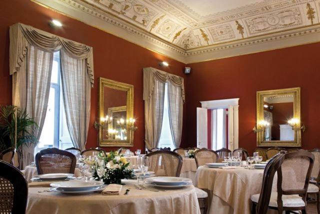 Parte do Napolit'amo Hotel Principe com várias mesas e cadeiras com taças, pratos e uma mesa com um arranjo de flor, há também duas grandes janelas com cortinas e um espelho quadrado com moldura dourada na parede do fundo, ilustrando post Hotéis em Nápoles.