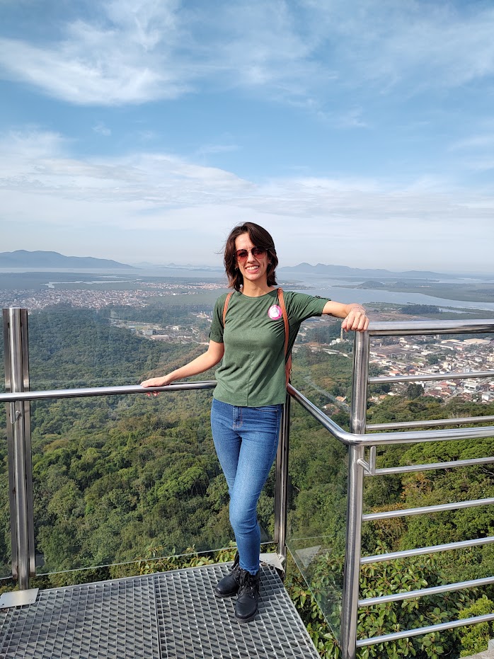 autora da postagem, uma mulher de cabelo curto e pele morena, está sorrindo e posando de pé para uma foto na grade do Mirante de Joinville. Atrás dela há toda a paisagem panorâmica.