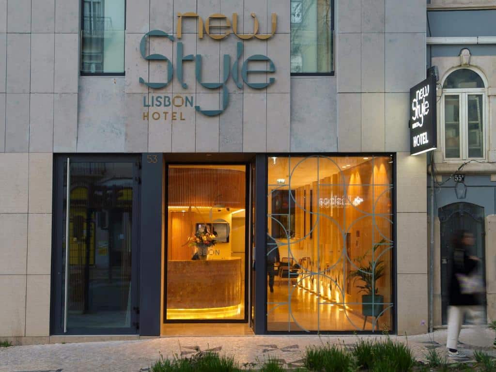 Entrada do  New Style Lisbon Hotel com uma porta de vidro com alguns desenhos geométricos, dentro há uma recepção iluminada com uma luz amarela, o prédio é cinza