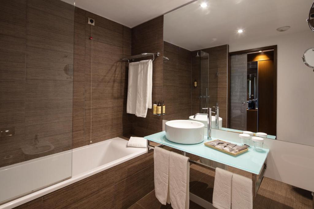 Banheiro do NEYA Lisboa Hotel com uma banheira do lado esquerdo, um espelho muito amplo sob a pia com uma cuba branca e diversos itens de higiene, além de toalhas brancas espelhadas pelo local, para representar hotéis baratos em Lisboa