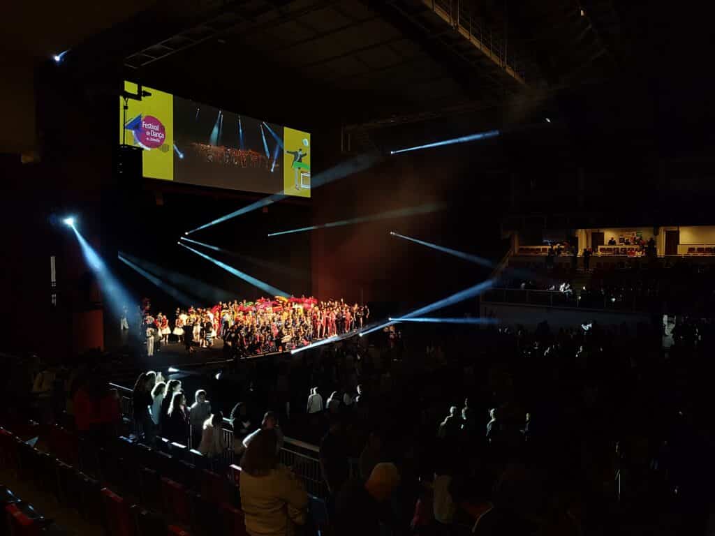 estádio de eventos do Festival de Dança de Joinville com um palco iluminado ao fundo, onde um aglomerado de pessoas está reunido, usando diferentes roupas coloridas. Todo o espaço está com as luzes apagadas, exceto no palco