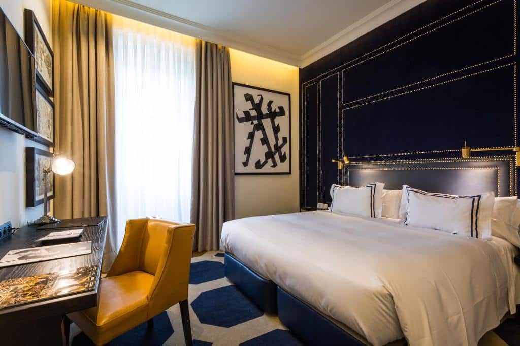 Quarto do Only YOU Boutique Hotel Madrid, uma das recomendações de hotéis românticos em Madri. A cama de casal com jogo de cama branco está encostado na parede azul e encara uma mesa de trabalho com luminária, cadeira e TV na parede acima. A parede ao lado tem uma grande janela com cortinas bege e brancas e um quadro ao lado.