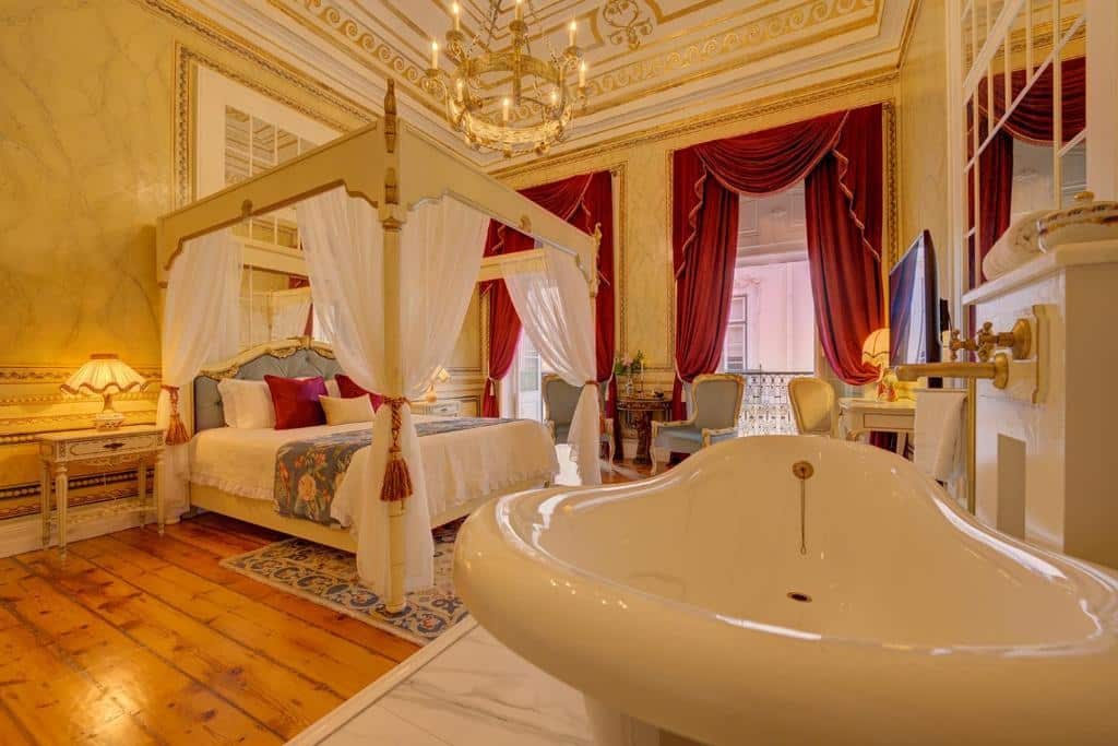 Quarto luxuoso do Palácio das Especiarias com duas sacadas com cortinas vermelhas, o ambiente inteiro remete a realeza, há um cama de casal com um bangalô com tecidos brancos, há um lustre dourado e também uma banheira de hidromassagem, para representar hotéis românticos em Lisboa