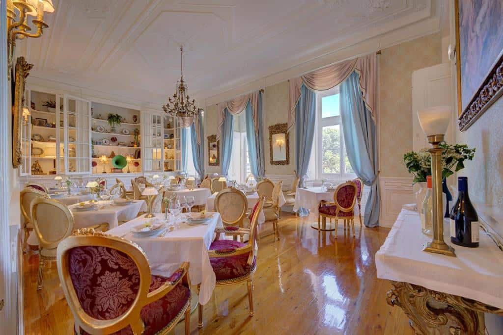 Salão de refeições do Palácio das Especiarias com muito luxo e com uma decoração que remete a realeza, com janelas com cortinas brancas e azul claro, há mesas quadradas e cadeiras estofadas em vermelho com dourado, chão que imita madeira e há também um lustre dourado no local