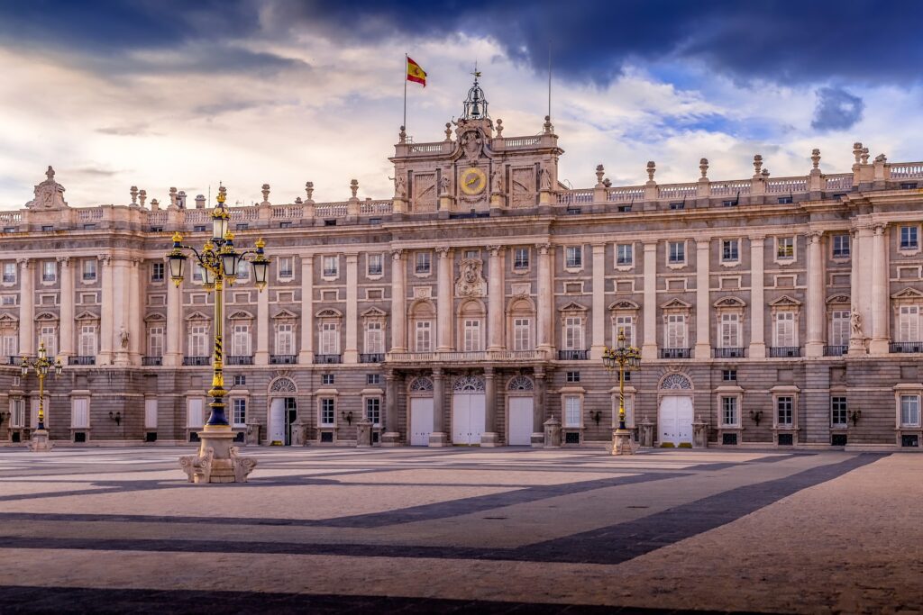 Palácio bege repleto de colunas e janelas. Há postes de luz na praça em frente, e uma bandeira da Espanha está tremulando ao vento perto do relógio que fica no centro do prédio. - Foto: ddzphoto via Pixabay