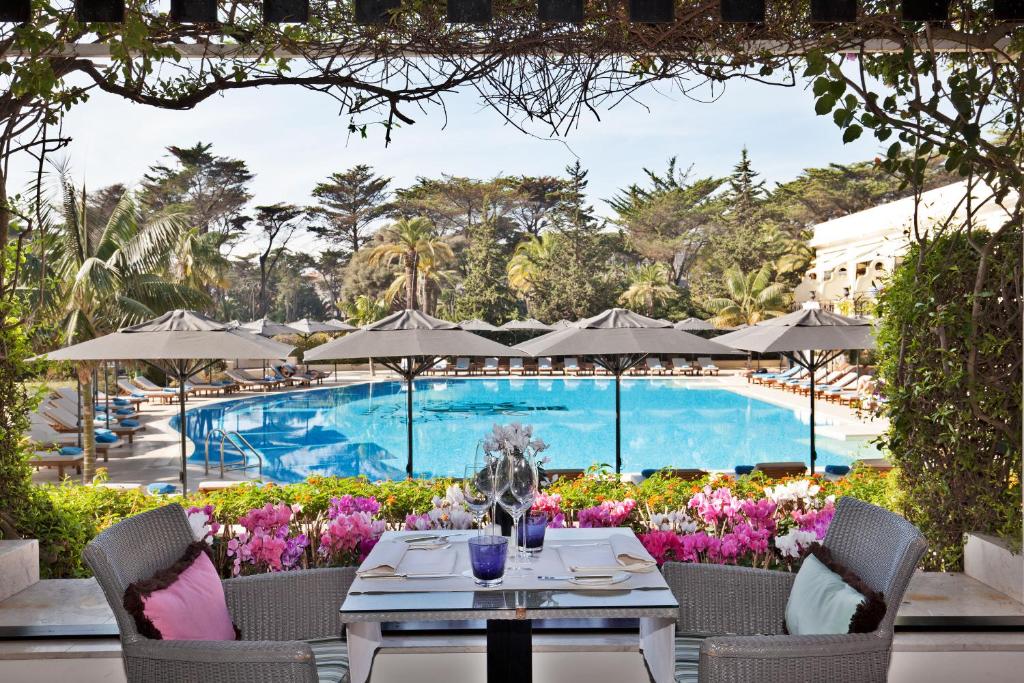 Piscina do Palácio Estoril Hotel, Golf & Wellness com muitas árvores e flores ao redor, e há também uma mesa quadrada com taças e velas em cima, e duas cadeiras com almofadas coloridas, para representar hotéis românticos em Lisboa