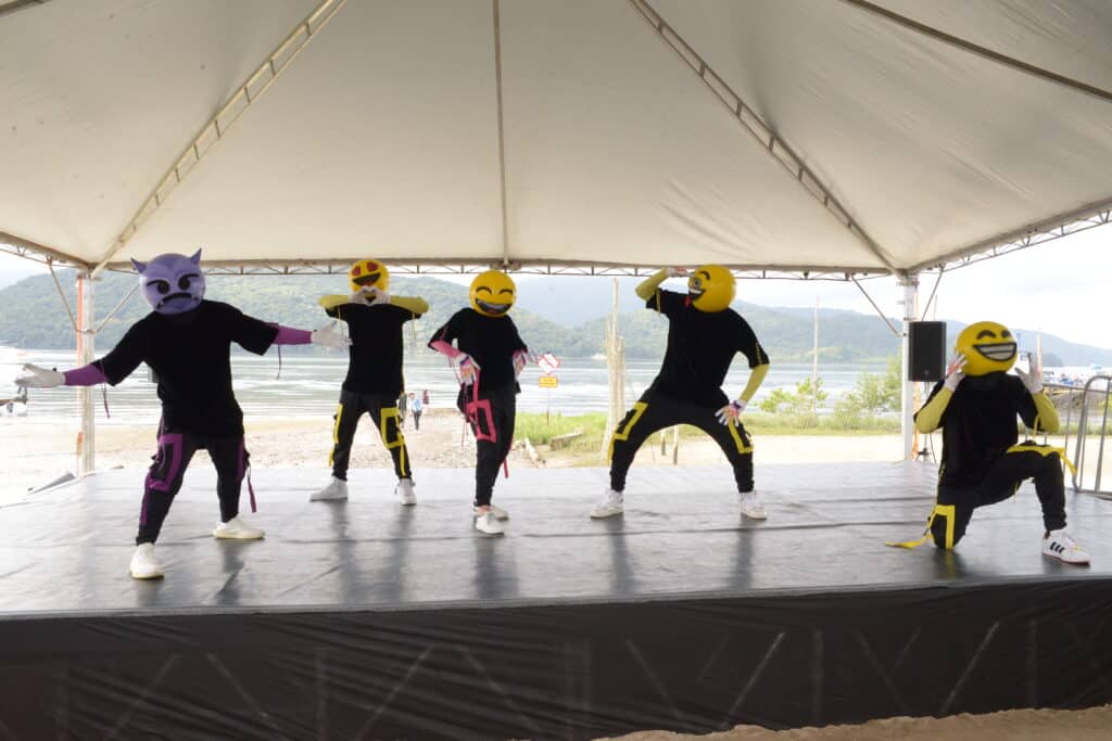 cinco pessoas vestidas de preto, fazendo poses e usando máscaras de emoji num palco aberto do festival de dança de joinville. Atrás do palco é possível enxergar um lago e algumas ilhas ao fundo, sob um dia claro.