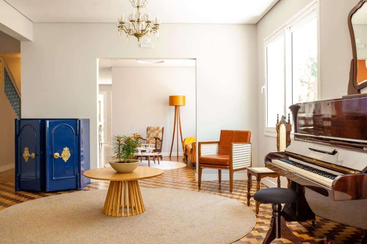 Recepção do Paranaguá Lodge. Um piano no lado direito, atrás uma poltrona. No lado esquerdo um armário azul. No centro da sala de recepção, uma mesa redonda e um lustre. Foto para ilustrar post sobre pousadas em Paranaguá.