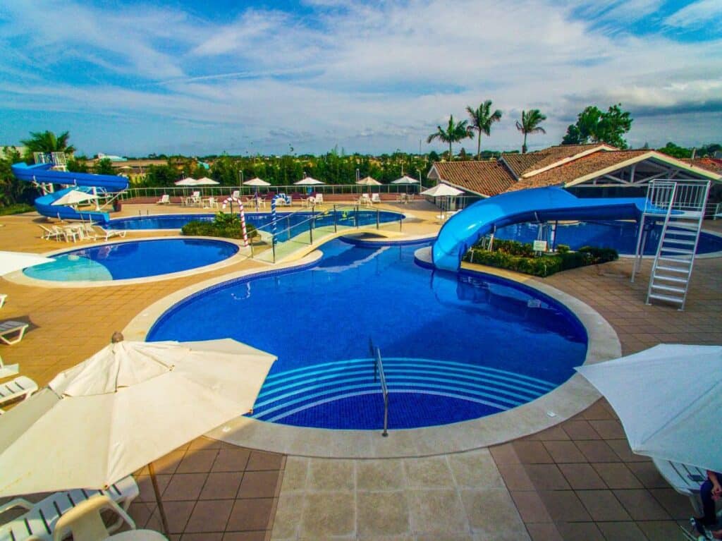 Piscina ao ar livre do Camboa Hotel Paranaguá. Quatro piscinas ligadas, em formato de círculo. No lado direito um escorregador pra piscina, no lado esquerdo um toboágua. Ao redor guarda-sóis e cadeiras de praia.