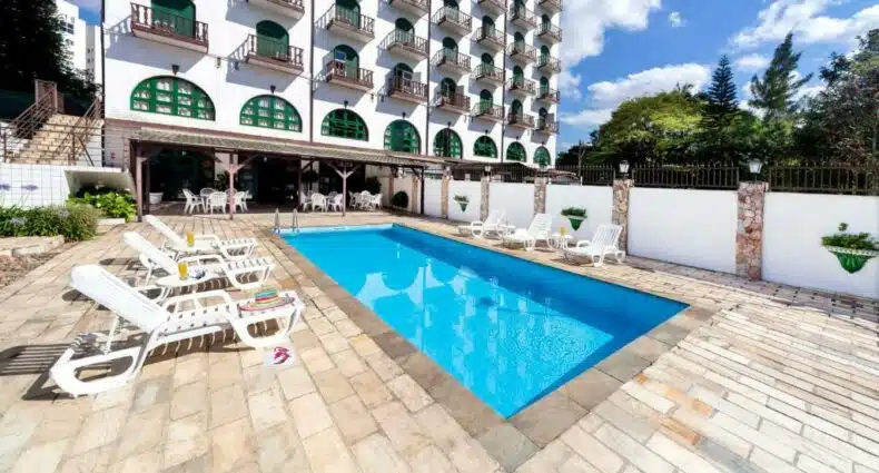 Piscina externa do Hotel Tannenhof em Joinville com espreguiçadeiras e mesas ao lado de um edifício de vários andares do hotel com varandas em um dia ensolarado. Representa uma das opções no post de hotéis em Joinville.