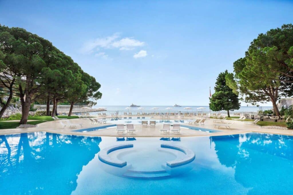 Piscina do Le Méridien Beach Plaza. Uma piscina redonda pequena atrás e outra grande na frente, ao ar livre. Ao redor cadeiras de tomar sol e árvores. No fundo, o mar.