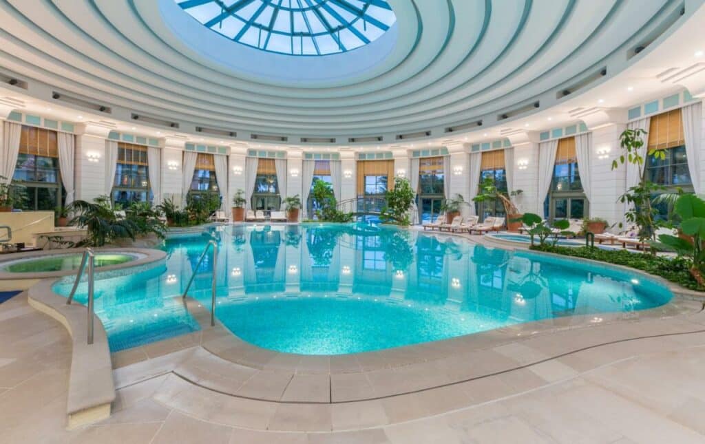 Piscina coberta do Monte-Carlo Bay Hotel & Resort. Uma piscina com banheira de hidromassagem dos dois lados. Cadeiras de tomar sol ao redor e plantas. Janelas na área com vista para a cidade.