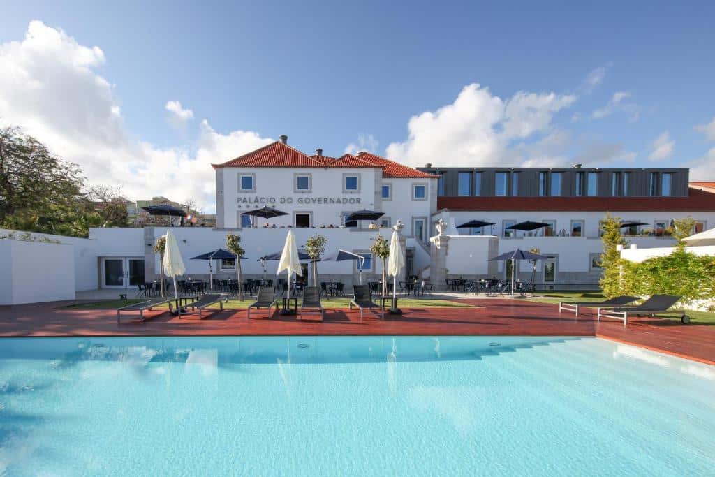 Ampla piscina do NAU Palacio do Governador com um deck de madeira ao redor com espreguiçadeiras e guarda-sóis brancos, para representar hotéis bem localizados em Lisboa