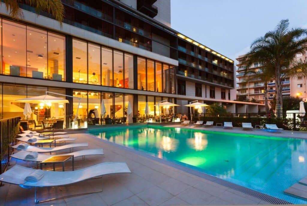 Piscina do Novotel Monte-Carlo. Uma piscina retangular iluminada, dos dois lados cadeira de tomar sol. No fundo, o hotel de vidro iluminado.