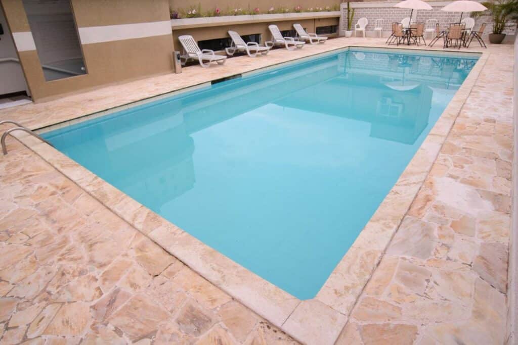Piscina do Porty Brasil Hotel. Uma piscina retangular, atrás mesas redondas com cadeiras e cadeira de tomar sol.