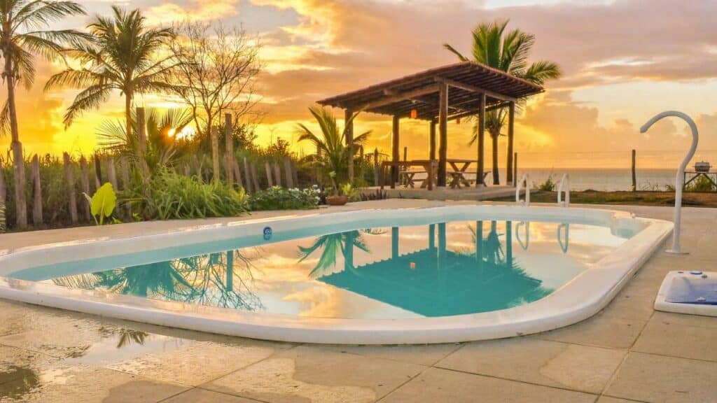 Piscina da Pousada Azul. Uma piscina na frente, atrás uma mesa de madeira com um teto demadeira. Do lado esquerdo da piscina um cercado de madeira e atrás mato. No fundo, a vista da piscina para o mar.