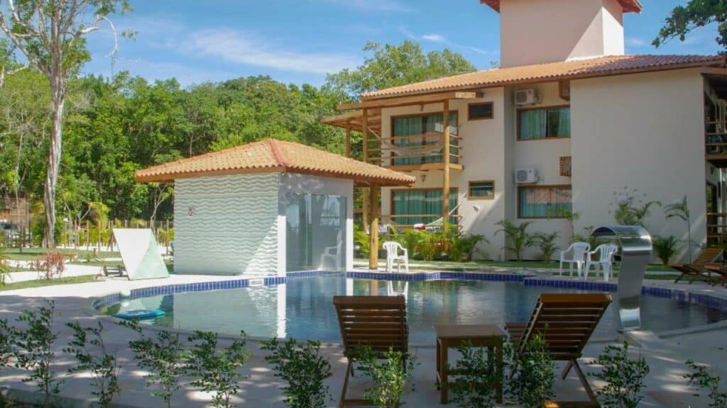 Piscina da Pousada Maré. Uma piscina redonda no meio, no lado direito uma cascata de água. Ao redor cadeiras de tomar sol. No lado esquerdo da piscina a sauna. Atrás a pousada.