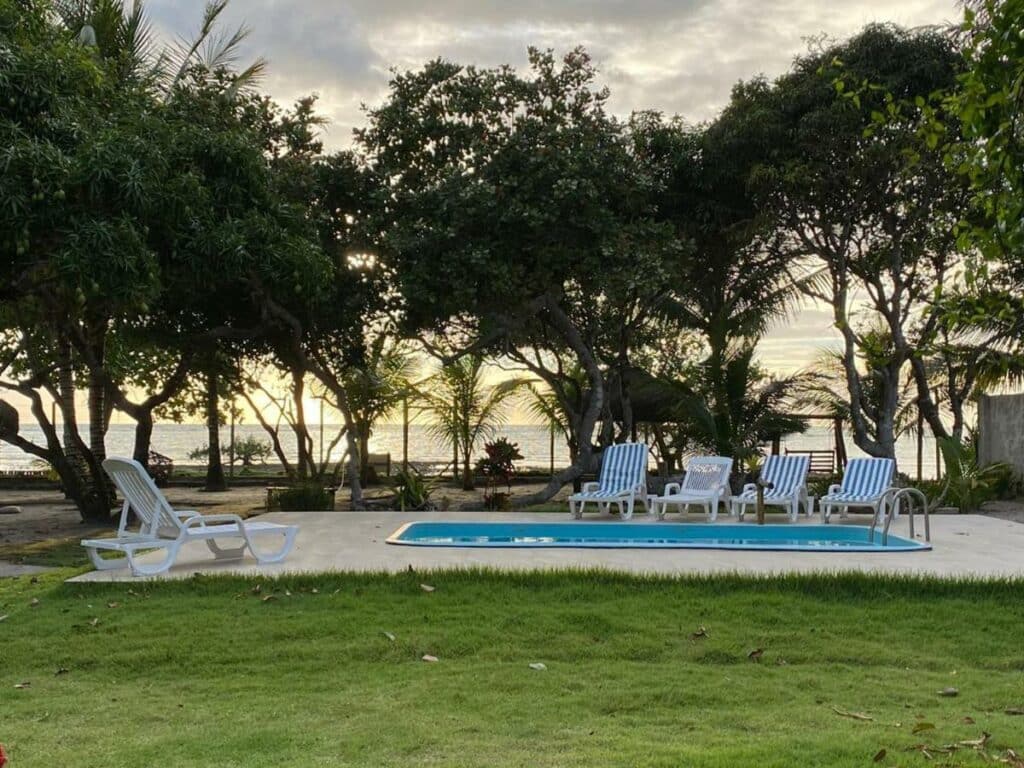 Área externa da Pousada São Francisco. Grama ao redor da piscina com cadeiras de praia. Atrás da piscina, árvores e a vista do mar.