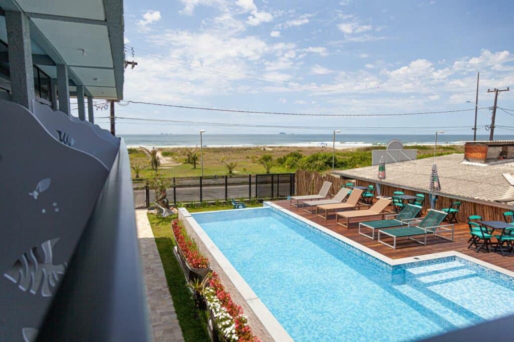 Piscina da Pousada Guaricana. Uma piscina no lado direito, com cadeiras de tomar sol. Do lado esquerdo a sacada da pousada. No fundo, a vista da praia.