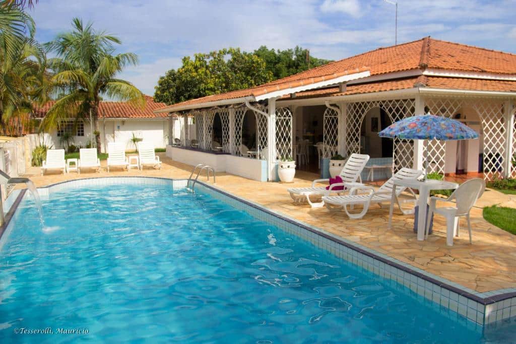 Imagem da piscina em Recanto Sereníssima, São Pedro. A piscina é longa e possui escada para acesso e chafariz. Ao redor do deck há espreguiçadeiras e brancas e uma mesa com guarda-sol.