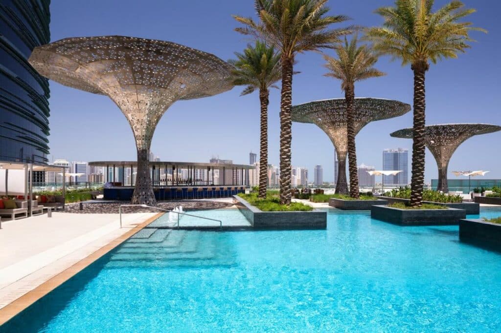 Piscina do Rosewood Abu Dhabi. Uma escada na piscina no lado esquerdo, a piscina no lado direito, com algumas palmeiras no meio. Do lado esquerdo o hotel. No fundo umas esculturas de palmeira e um bar.