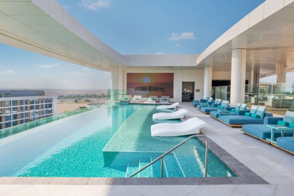 Piscina do The WB Abu Dhabi, Curio Collection By Hilton. Uma piscina de borda infinita no terraço. Do lado esquerdo a vista para a cidade, do lado direito cadeiras de tomar sol fora e dentro da piscina. Na frente, a escada da piscina.