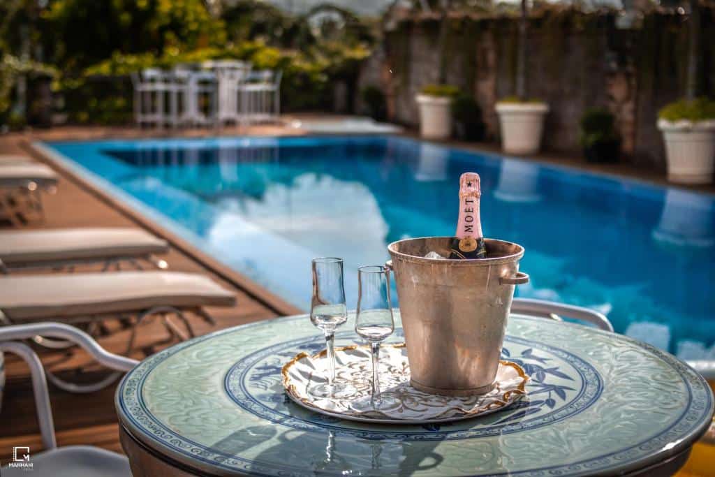Imagem tirada na piscina da pousada Villa Maria Pousada de Charme. Vemos uma mesa com um balde com champagne e duas taças e mais espreguiçadeiras ao redor da piscina.
