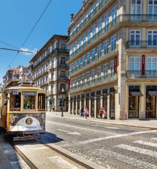 Rua no Porto com edifícios em volta e um bondinho do lado esquerdo da imagem durante o dia. Representa onde ficar no Porto.