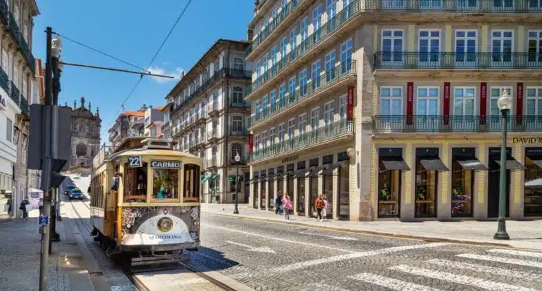 Rua no Porto com edifícios em volta e um bondinho do lado esquerdo da imagem durante o dia. Representa onde ficar no Porto.